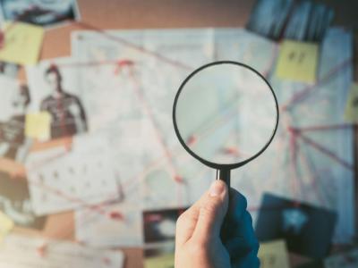 person holding a magnifying glass to a board full of little notes of clues to a mystery